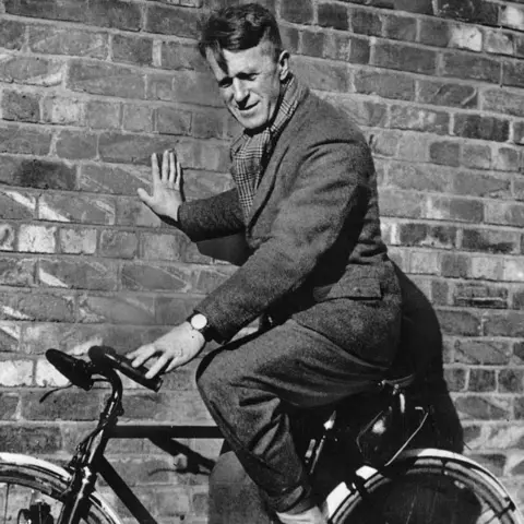Hulton Archive/Getty Images A black and white picture of a man wearing trousers and a coat, sitting on a bike and leaning against a brick wall. He also has a scarf around his neck.