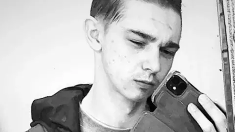 Facebook A black and white image of Scott Jeff. He has short hair and is wearing a top with a dark hood. He is holding a phone and has a serious expression on his face.