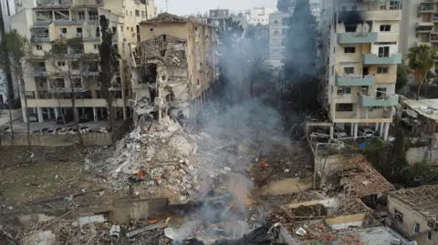 A landscape image of a damaged residential area. Smoke rises from the floor while buildings have collapsed into rubble.