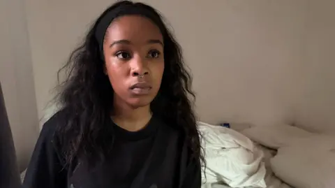 Isabel Rose is seen wearing a black top and a small nose ring, sits facing the camera indoors.