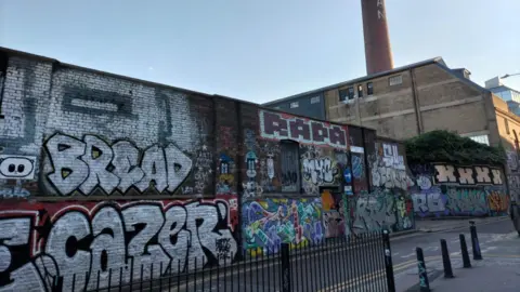 LDRS Graffiti-covered walls at the Truman Brewery site in Brick Lane, with the brewery's distinctive chimney rising above industrial buildings in the background.