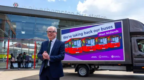 Parker is stood outside Wolverhampton bus station with a van advertising the new franchise system. It depicts red buses with a strapline above that says: We're taking back our buses