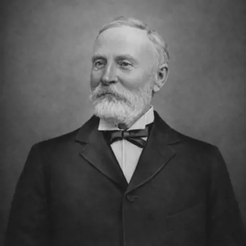 Chris Middleton A black-and-white photograph of a distinguished-looking man with short grey hair and a white beard. He is wearing a black, buttoned jacket over a starched white shirt and a black bow-tie.