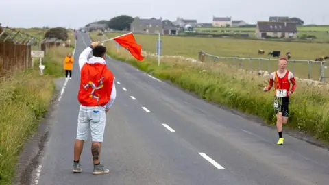 Vannin Photos Corrin Leeming wears a red and black lycra running top and shorts, and sunglasses with yellow trainers. He is running on a country road with lots of fields nearby, with some cows in the distance. A man stands on the opposite side of the grey road and waves a Manx flag. The spectator also has a red Manx flag draped over his shoulders.