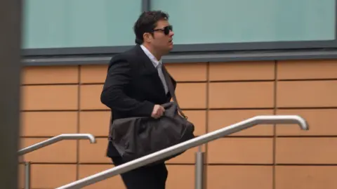 BBC A man with black hair walking into court with a duffle bag around his shoulder. He's wearing a black suit and white shirt. The building behind him is orange/brown brick. 