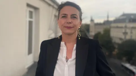 Os elodes de Elodi Telilian-Homan são fotografados na varanda em Paris. Ela olha para a câmera e usa o casaco branco e o casaco preto com orelhas douradas. 