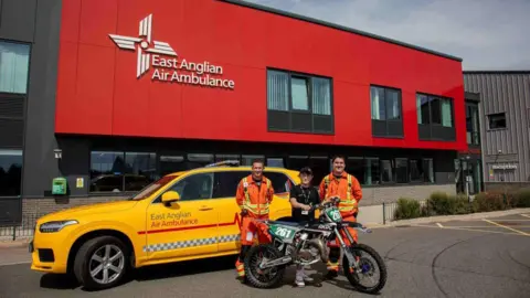 East Anglian Air Ambulance Jaden, 13, dressed in black stands with his motocross bike in front of him. He is standing between two air ambulance crew members in orange flightsuits. They are all next to a yellow air ambulance charity car outside the base in Norwich.