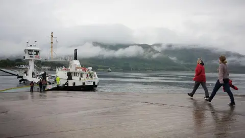 The Corran Ferry is a white and back boat. It is berthed at a slipway. Two women walk down the slipway towards the ferry. Mist hangs on the hills on the opposite shore.