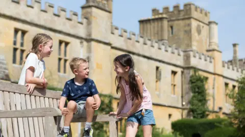 Sudeley Castle and Gardens Three young children - a boy and two girls are on a bench outside a castle, which has a tower and battlements against a blue sky. The children are in summer shorts and t-shirts and are looking at each other and smiling.