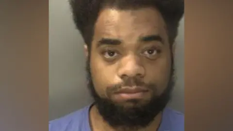 A man who has dark hair curly hair and a dark beard is photographed in front of a neutral background. He has a light blue t-shirt on and appears expressionless.