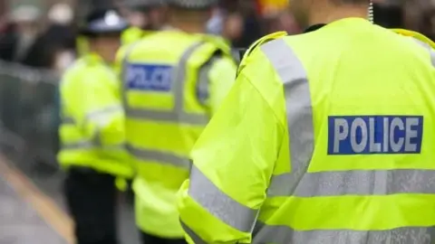 The back of a police officer who is wearing a yellow hi-vis jacket with silver reflective strips. It says "police" on the back. In the background are two other officers, wearing the same jackets as well as black police hats.