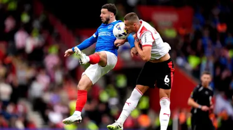 Portsmouth's Conor Chaplin battles for possession of the ball in the air with Southampton's Taylor Harwood-Bellis. Both men are captured vying for the ball in midair with Harwood-Bellis managing to get his head on it.