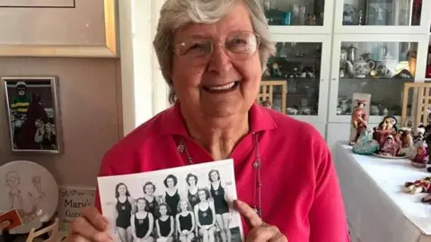 Mary Black when she was in her 80s. She has short grey hair and glasses and is wearing a pink jumper. She is holding an old photograph of her synchronised swimming team from when she was a teenager. There are figurines and ornaments in the background.
