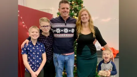 A family of five stands together in front of a Christmas tree in a group hug. On the left side, under their father's arm, are two blonde boys. In the centre is a man with short grey hair wearing a navy blue Christmas jumper, to his right is a woman with long strawberry blonde hair wearing a black top and green skirt, and to her right is a small boy with blonde hair and an excited expression.