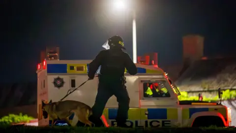 A PSNI dog handler stands infront of a police land rover. He is wearing a helmet and protective clothing. the night sky is dark, with a street lamp in the middle of the image. Behind the police land rover is a row of houses. 