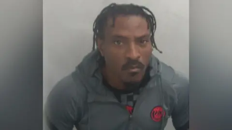 Essex Police A police custody photo of Temitope Adeyinka. He has a stern look on his face. He has braided black hair, a moustache and a beard. He is wearing a grey hoodie with a grey and black chequered top underneath.