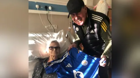 Janet Wheatley, lying in a hospital style bed, with Alan Birchenall standing next to her and holding a blue Leicester City football shirt