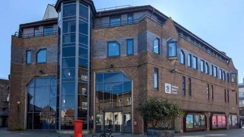 A large brown local authority building with large windows in the front. It has a red post box and two bicycles in front of it. 