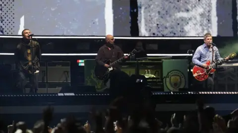 PA Media Oasis are on stage at the Principality Stadium, Cardiff, as the band's long-awaited reunion tour kicks off in Wales. Both Liam and Noel Gallagher are singing into the microphones as fans in front of them wave their arms in in the air.