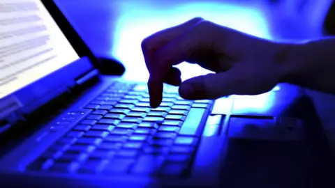 A person's hand is seen typing on a laptop keyboard in a dark room in a stock image. 