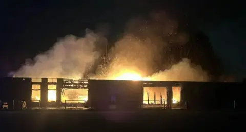 Flames and smoke can be seen rising from the gutted rugby club building. It is night time.