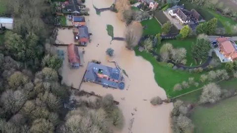 Aerial view of six homes being inundated with brown floodwater. On the left of the homes are dense trees.