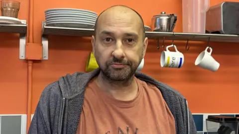A photo of a man standing in a kitchen with cups hanging up in the background. He is wearing an orange t-shirt and grey jacket