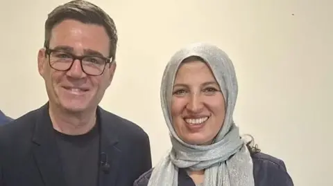 A man in a dark suit and T shirt, with dark-framed glasses, stands with a woman wearing a silver headscarf.