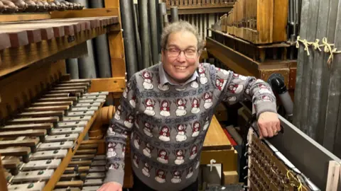 Mr Martin smiling to camera in a Christmas snowman jumper, at the back of the organ showing all the mechanics.