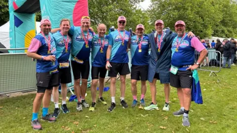 Huw Jones The eight fathers who took part in the challenge, wearing T-shirts, dark shorts and pink hats and medals, standing on grass.