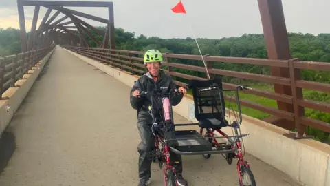 Supplied Nahla Summers is sitting on one seat of the side-by-side tandem bike and smiling brightly at the camera. She is wearing a dark, waterproof outfit and a bright yellow/green helmet. There is a red flag erected on the bike. She is on a brown metal bridge with a archway design overhead that resembles a spiralling vortex.