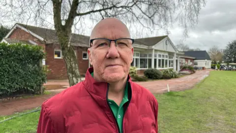 A man with a bald head is standing on a golf course and looking into the camera. He has black rimmed glasses and is wearing a red-zipped jacket. Behind him is the clubhouse.