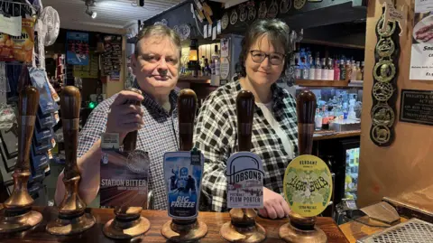 A man and a woman are standing behind a bar inside a pub. There are beer taps across the counter. The man has grey hair and is wearing a checked shirt. The woman has short broken hair and is wearing glasses.