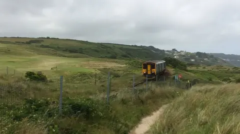 A two-carriage GWR train on a track among fields with a gravel footpath besides it on a slightly overcast day.