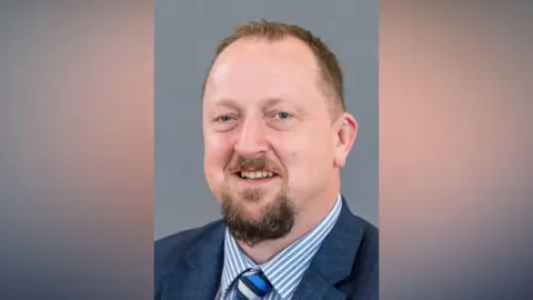 Colin Kennedy looking at the camera. He has brown hair and a beard. He is wearing a navy suit jacket, striped blue and white shirt and a blue and navy striped tie.