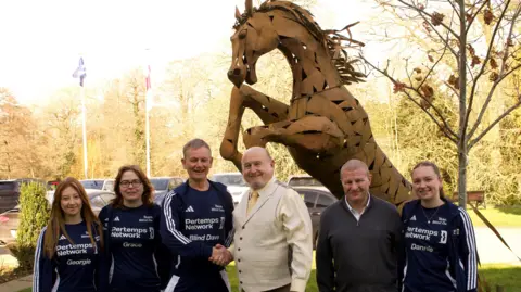 Six people, three men and three women, stood in front of a large bronze horse sculpture. The two men in the middle, one in a blue running kit with Blind Dave written on it and one in a cream waistcoat suit are shaking hands.