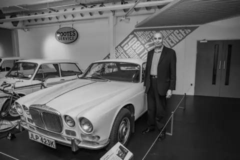 Günter and Christine Starke Harbhajan Singh Pangli stands next to a Daimler at Coventry Transport Museum. He is wearing a dark suit with lighter shirt. 