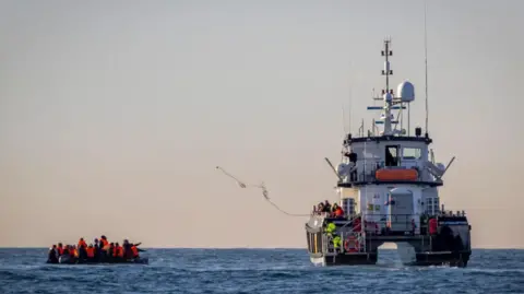 A group of migrants are in a boat in the Channel closely followed by a border patrol vessel