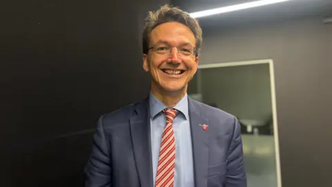 Kirsten Morel is dressed in formal attire, wearing a navy blue suit jacket, a light blue shirt, and a red-and-white striped tie. The background features a dark wall with a bright horizontal light fixture and a reflective surface, suggesting an indoor professional or studio environment
