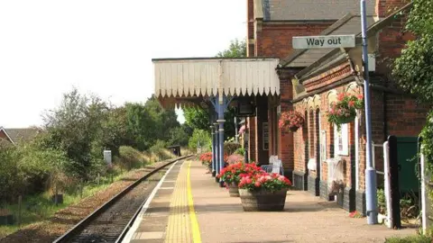 Evelyn Simak/Geograph A railway station on a sunny day can be seen, with flowers blooming in planters on the platform and a sign directing passengers to the way out.