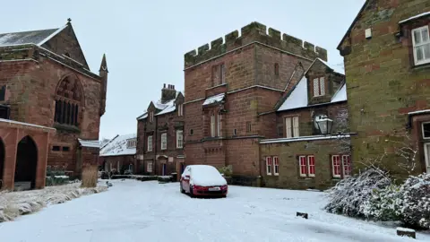 Carlisle Cathedral covered in a sprinkling of snow, which has settled on ledges, rooves, bushes and the top of a Victorian-style light. There is snow on the road and a thicker layer on a red car.