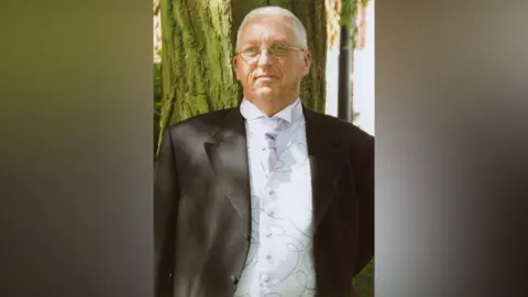 Family photograph Peter Moody dressed in formal wedding attire. He has a black suit and a white waistcoat, shirt and tie. He has grey hair and glasses and is standing in front of a tree.