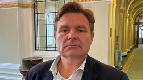 Council leader John Cotton pictured with a blank expression on his face. He has light brown hair which is styled in a quiff. He is wearing a dotted white shirt underneath a blue navy blazer