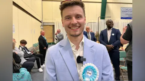 Nicola Haseler/BBC Liam Morrell Phillips smiles at the camera wearing his Reform white and blue badge on a pale blue suit. He has sunglasses pushed back on his head. Other people stand around behind him. 