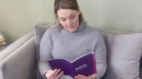 A woman wearing a grey jumper and sitting on a grey sofa. She has brown hair tied back in a ponytail. She is reading a purple booklet bearing the title 'Staying safe, finding hope'.