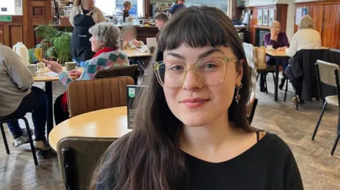 Ava Lewis is at a table in a cafe. In the background you can see tables and chairs with customers sitting at them. Ava is wearing a black top and glasses. 