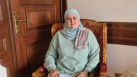 Caroline Bazzi/Jinha Agency Hania is sitting in a chair, smiling at the camera. She is wearing a pastel green top and a pastel blue headscarf. 