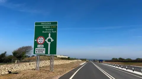 A picture from the side of the road. There is a green sign which shows a roundabout ahead and the arrow pointing up says 'Bodmin and Victoria Business Park" at the bottom of the sign it says "Padstow B3274". The side of the road there is a small stone wall and dirt and grass.