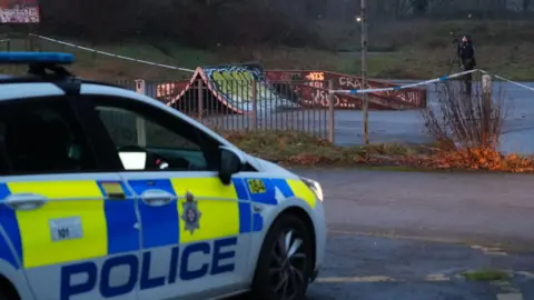 A skate park at dusk. The equipment is heavily graffitied. Police tape cordons off the area and a police photographer can be seen taking pictures. A police car is in the foreground. 