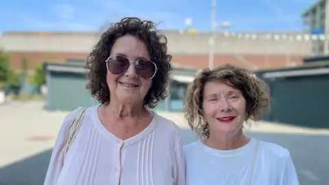 Laura Foster/BBC June Smith, wearing a white top and shades is standing in Basildon town centre with Teresa Smith standing next to her.
